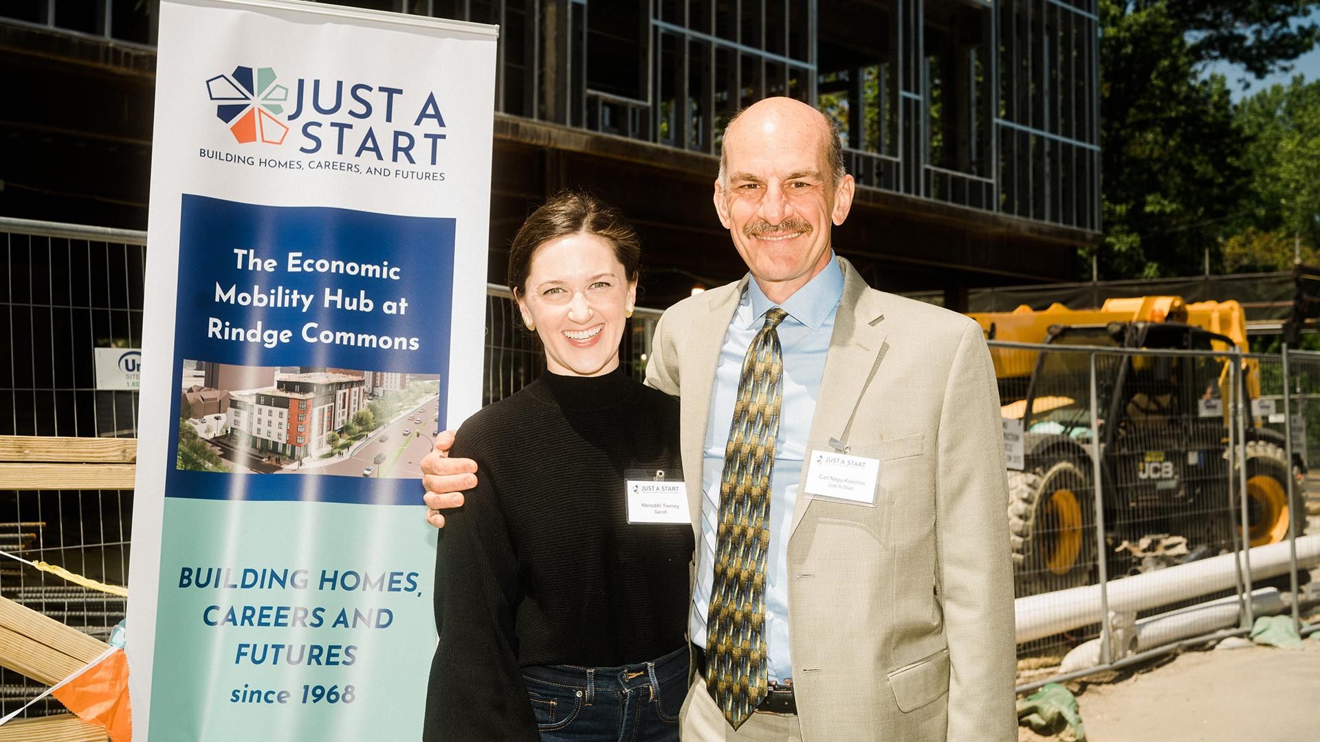 Two individuals stand side by side in front of a construction site. Meredith Tierney, on the left, wears a black top and jeans. A member of Just-A-Start, on the right, wears a light-colored suit with a patterned tie and has their arm around Meredith’s shoulder. Both wear name tags. A banner nearby reads “JUST A START – BUILDING HOMES, CAREERS AND FUTURES – The Economic Mobility Hub at Rindge Commons.