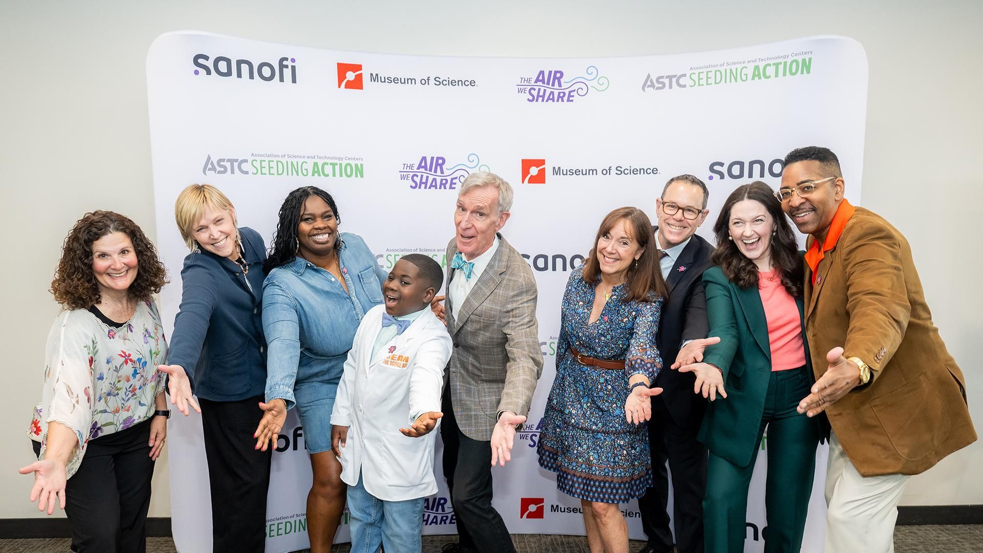 A celebratory group photo captures the diverse team behind "The Air We Share" educational program at the Museum of Science in Boston. Sanofi team members Erica Ferry, Barb Short, Tasha Wright, Diana Blankman, Dr. Michael Greenberg, Meredith Tierney and Phillip St. James stand together with Sean the Science Kid and Bill Nye, all smiling in front of a professional event backdrop prominently displaying "THE AIR WE SHARE" in large text, along with logos from ASTC Seeding Action, the Museum of Science, and the Association of Science and Technology Centers. The backdrop's branding emphasizes the collaborative nature of this initiative focused on connecting environmental conditions with respiratory health education and STEM literacy.