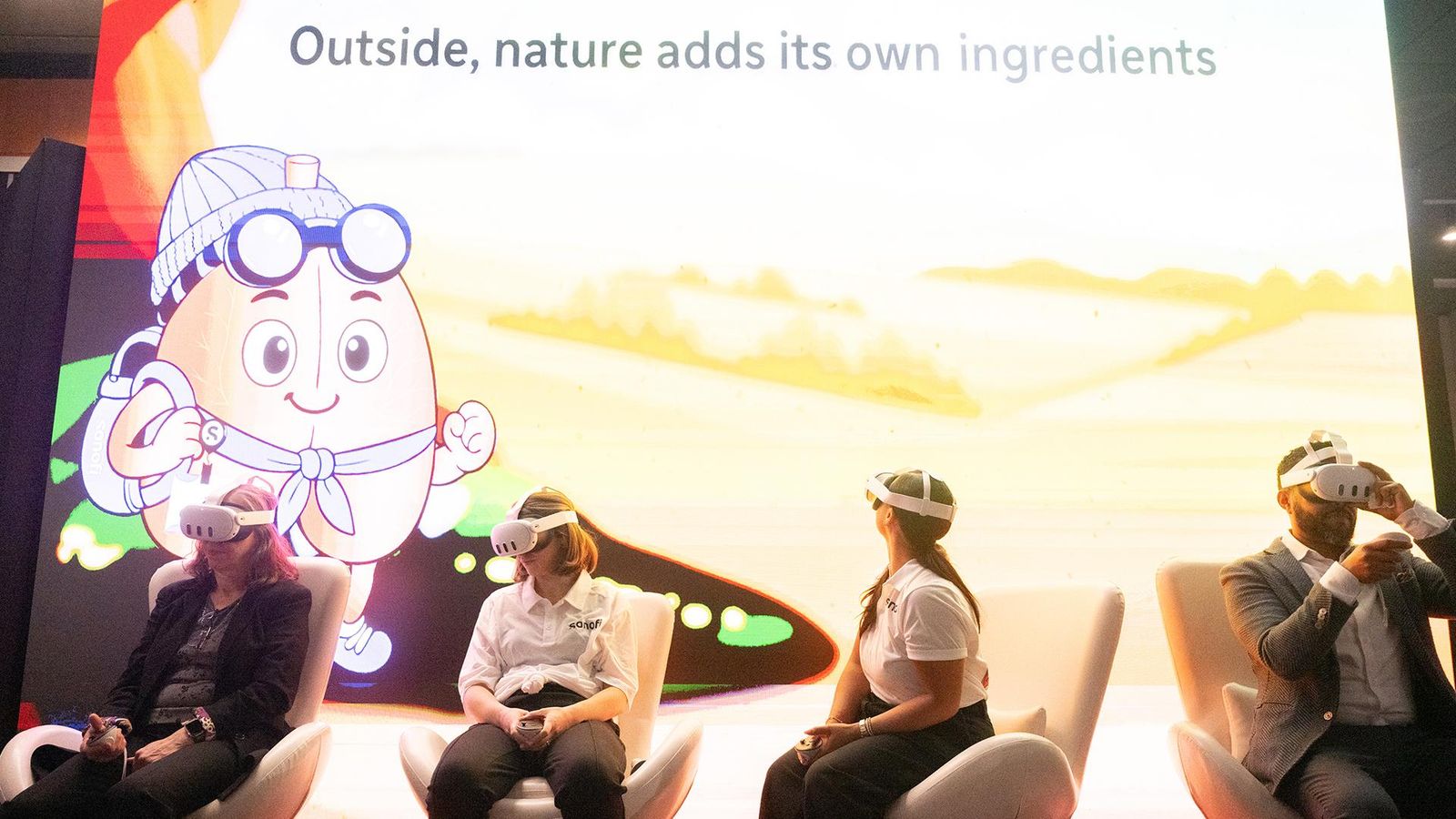 Four participants — including two Sanofi staff members in branded white shirts, one attendee in a dark jacket, and one in a suit and tie — sit side by side in white chairs, each wearing virtual reality headsets and holding VR controllers as they engage with "The Air We Share" immersive experience at the Museum of Science in Boston. The large display screen behind them features a vibrant, animated scene with a cartoon character dressed in goggles and a wide-brimmed hat, standing on a sunlit green field with a warm yellow horizon. The screen's headline reads "Outside, nature adds its own ingredients," reinforcing the exhibit's focus on how outdoor environmental conditions impact respiratory health. The Sanofi logo is prominently displayed, reflecting the company's sponsorship of this science education initiative.