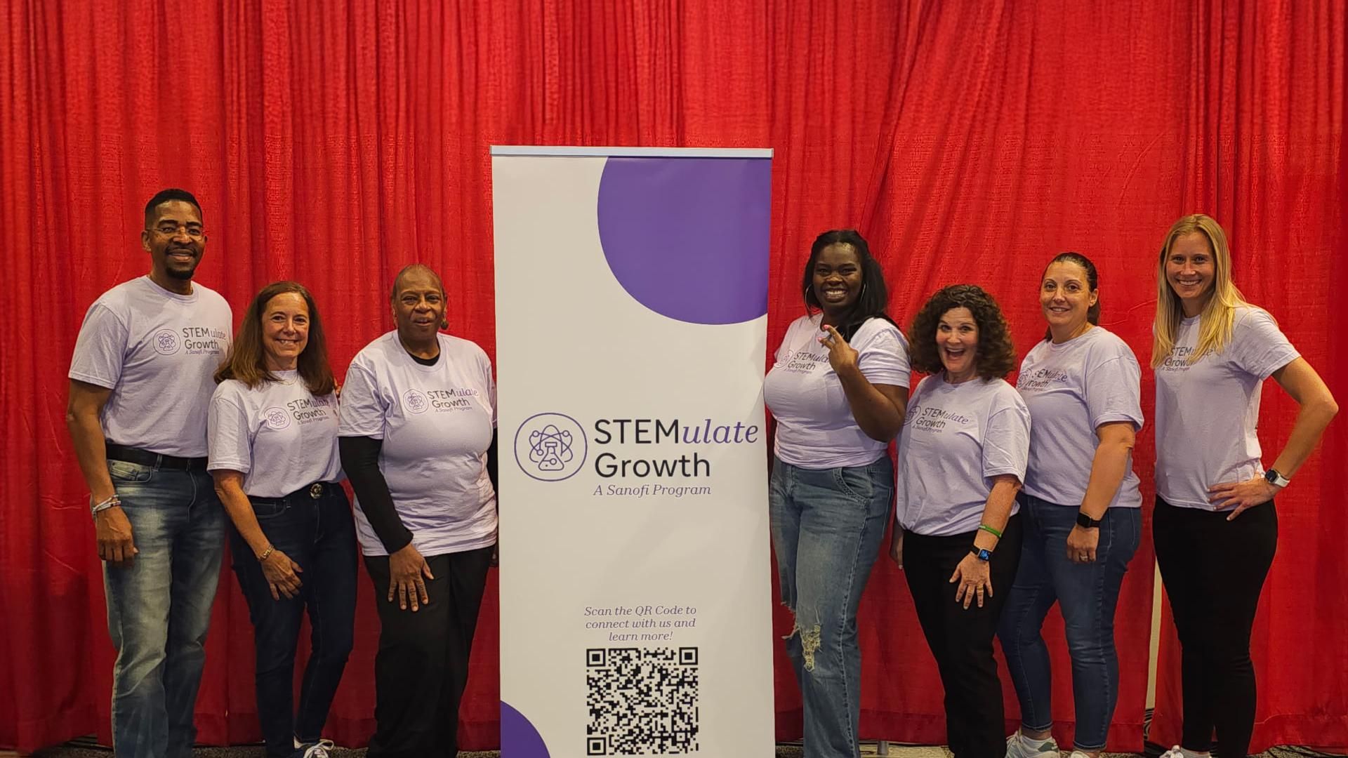 Eight Sanofi team members wearing matching light gray "STEMulate Growth" t-shirts smile and pose together in front of a red backdrop featuring a "STEMulate Growth – A Sanofi Program" sign at the Association of Science and Technology Centers event announcing the launch of The Air We Share. 
