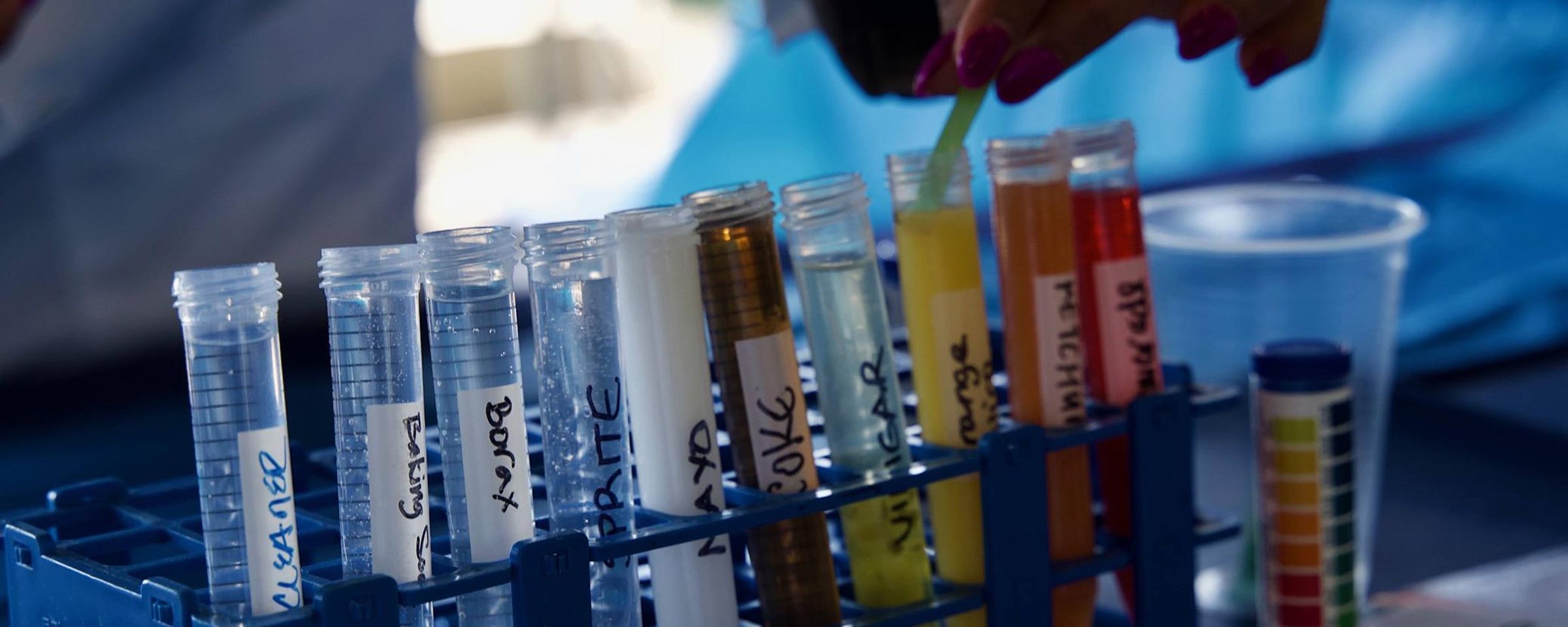 A close-up image of a student’s hand holding a strip above several test tubes filled with colorful liquids, arranged in a blue rack during a STEM education event.