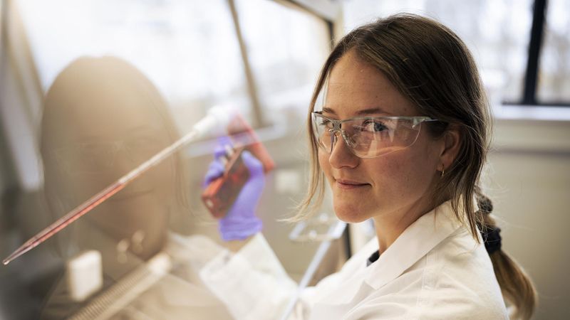 Layla Bral looks pointedly at the camera as she works under a fume hood moving red liquid between vials, wearing lab gear.