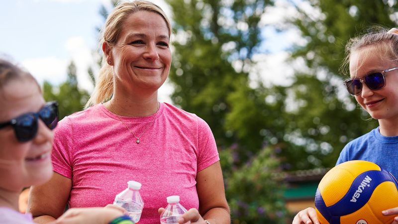 Caroline Blechschmidt, standing with 2 kids outside by some trees, holding 2 water bottles and wearing a pink t-shirt.