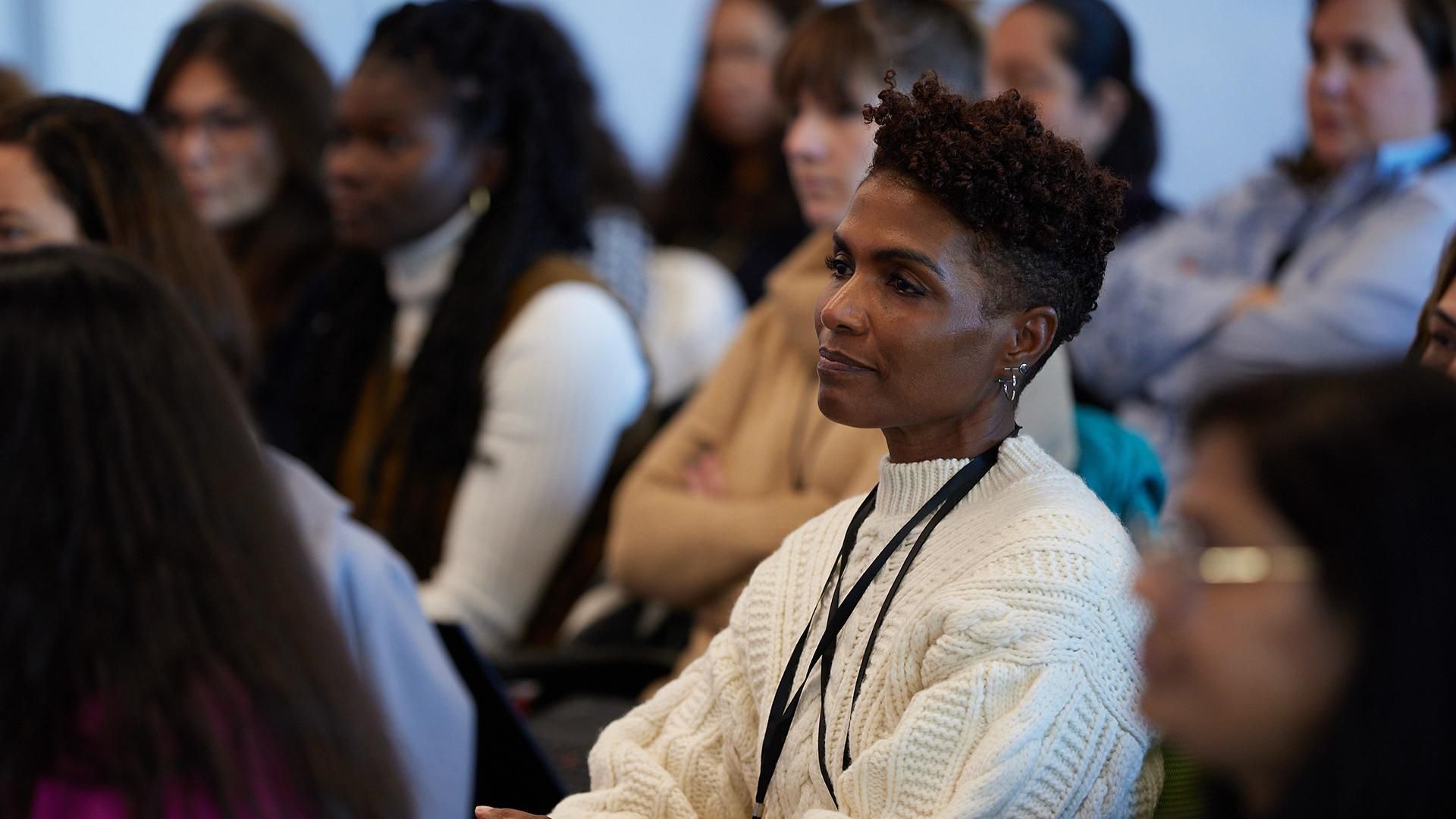 Jennie McGregor, wearing a white sweater and silver earrings with short dark hair and shaved sides, sits smiling in the audience.