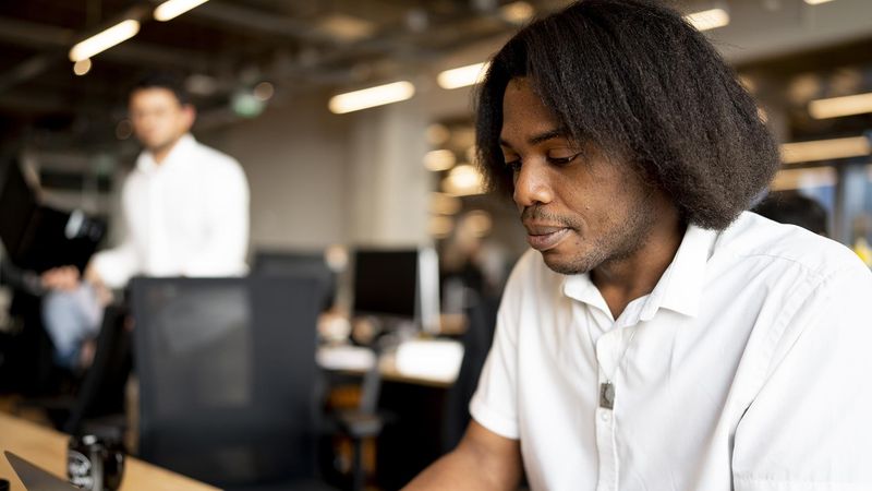 David Biteghe works on his laptop in a shared workspace wearing a white shirt and has coarse neck length black hair.