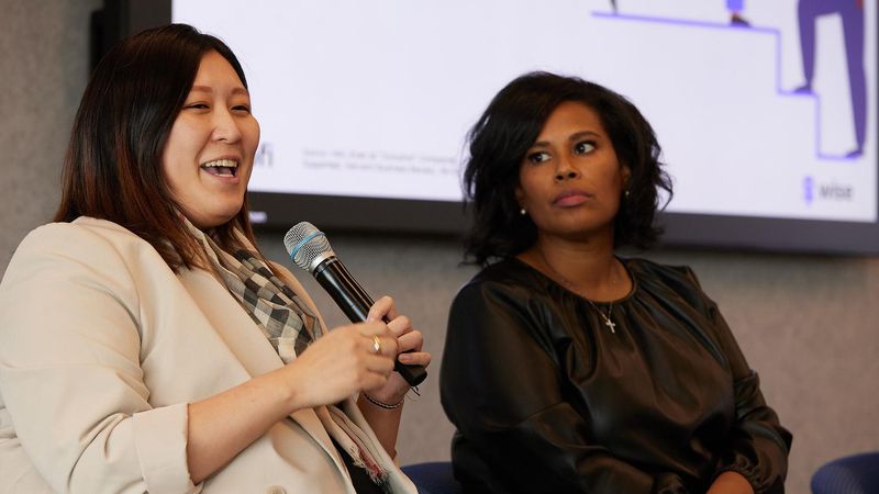 Jennie Jung (left), sits on stage speaking into a mic wearing a white suit, with Tanisha Sullivan (right) listening and waiting to speak.