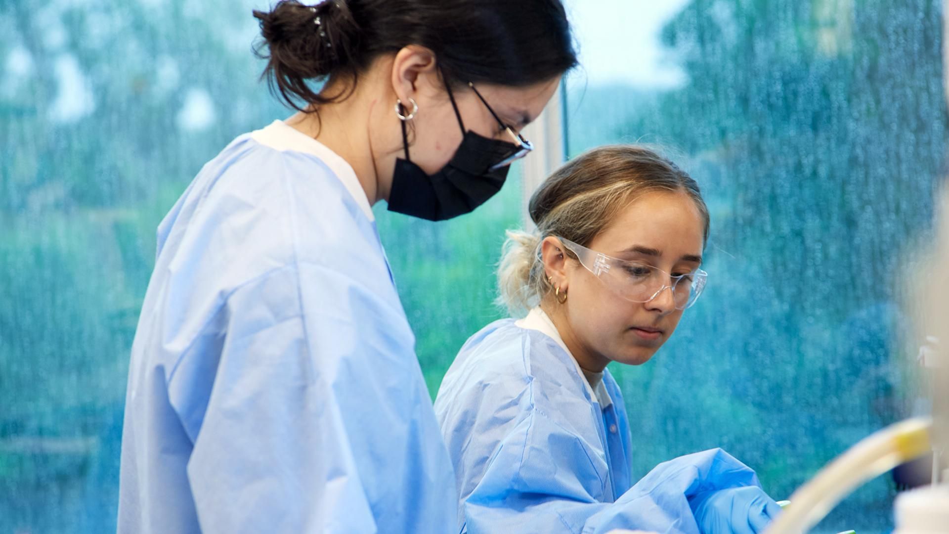 Abigail Leavitt and Kara Gilbert, wearing gloves, glasses, and blue lab outfits, moving vials around.