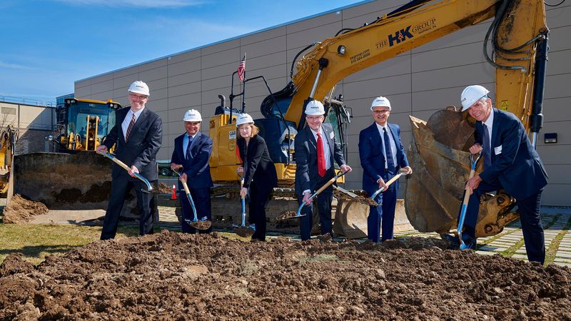 Six Sanofi executives and officials in business attire and white hard hats hold ceremonial shovels during the groundbreaking ceremony for Sanofi's new sustainable pandemic flu vaccine production facility in Swiftwater. An HK Group excavator with an American flag stands ready behind the group, with the facility building visible in the background under a clear blue sky.