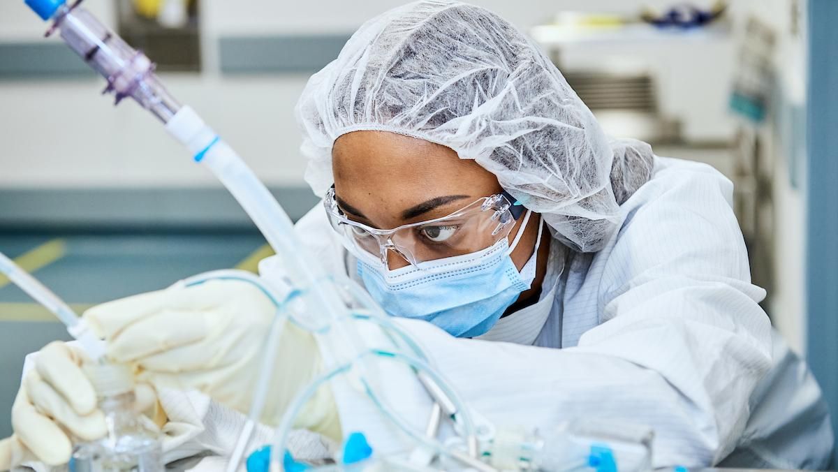 A laboratory worker wearing a hair cover, safety glasses, mask, and gloves closely adjusts equipment in a manufacturing lab.