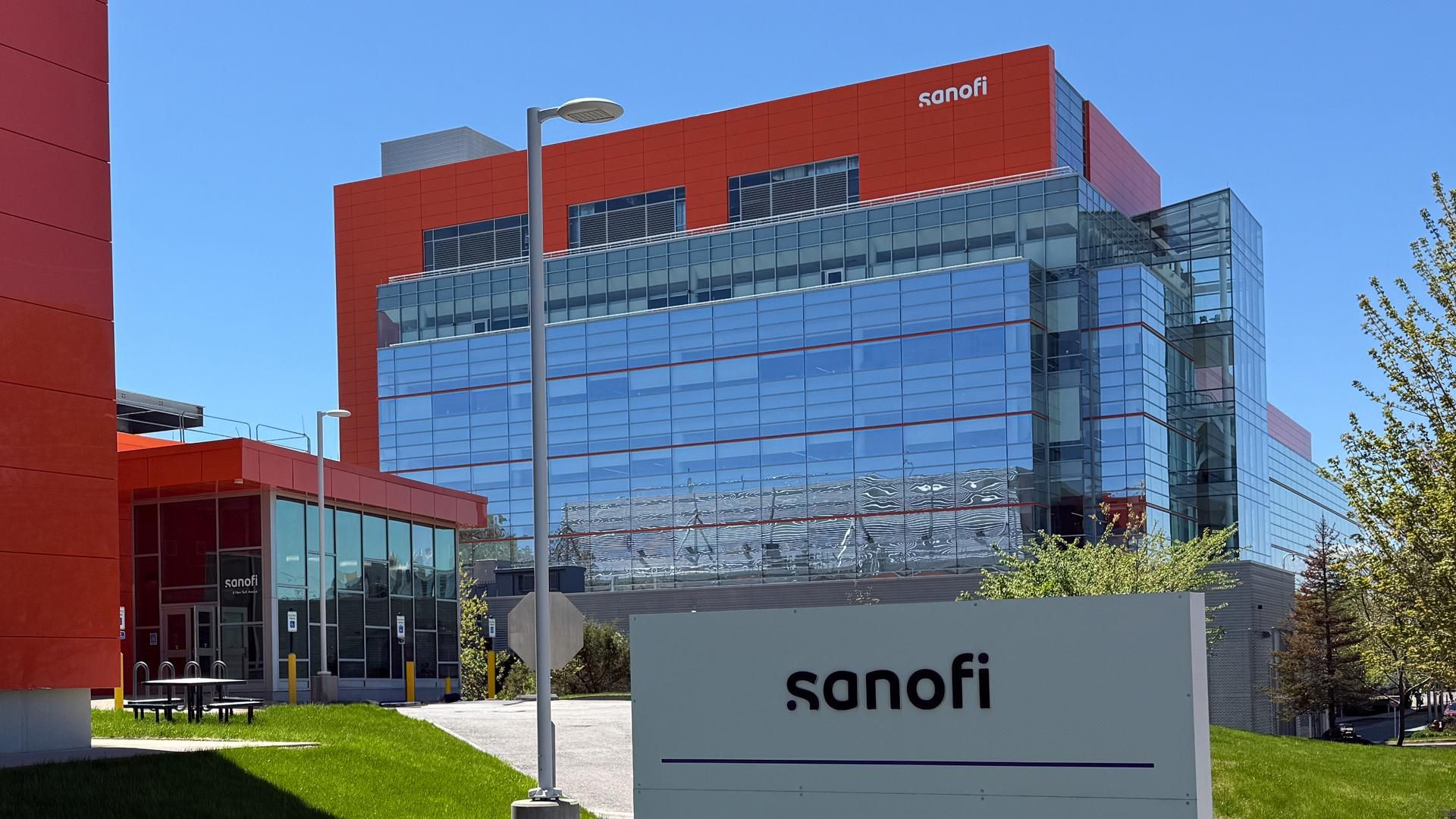 A modern multi-story Sanofi facility with a contemporary red and glass façade displaying the Sanofi name in white lettering. The building is surrounded by neatly trimmed grass, trees, and a picnic area, with a clear blue sky in the background.